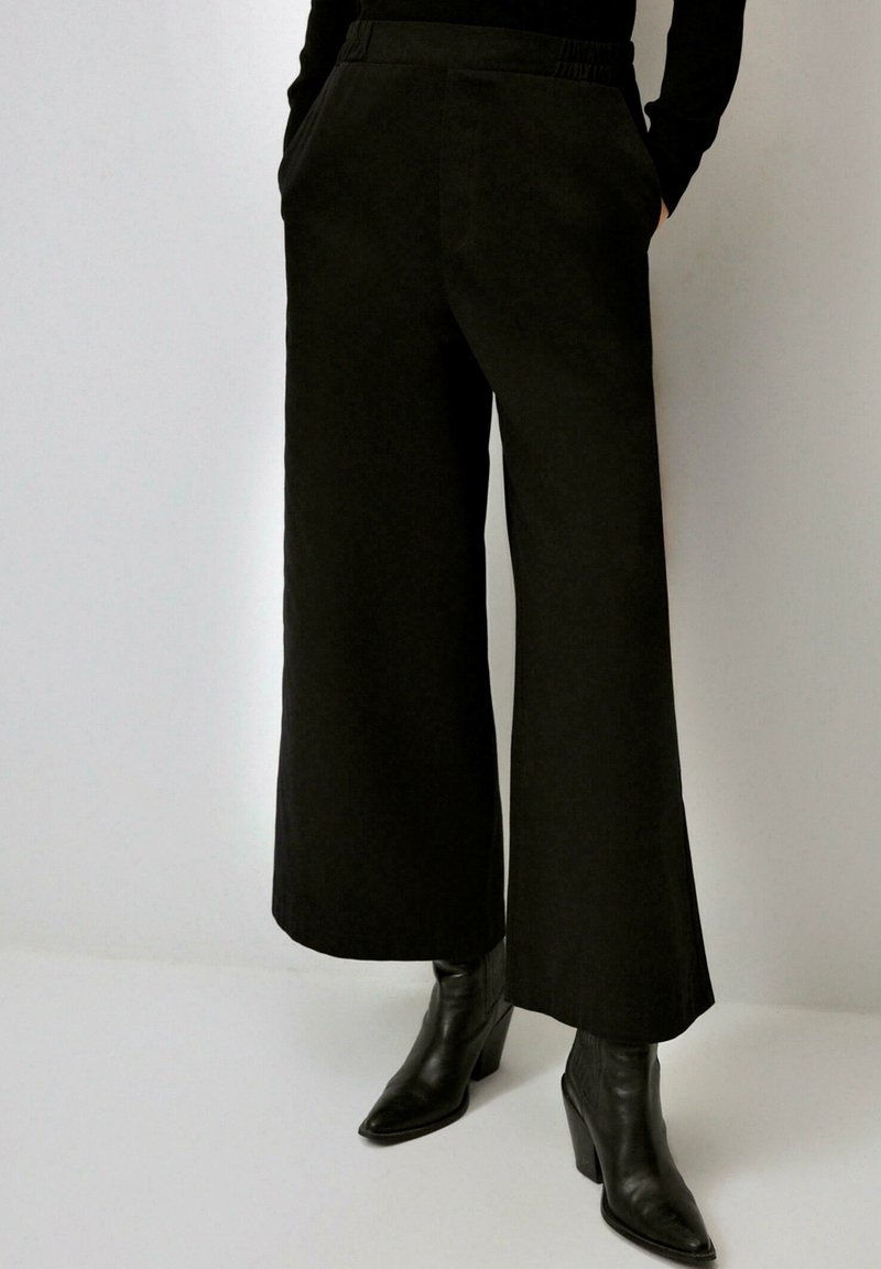 Pantalons larges noirs avec une taille élastique, des poches latérales et un tissu lisse ; associés à des bottines noires pointues.