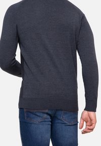 Hombre que lleva un suéter de manga larga gris oscuro y jeans azules, mostrado de espaldas con un brazo ligeramente doblado.