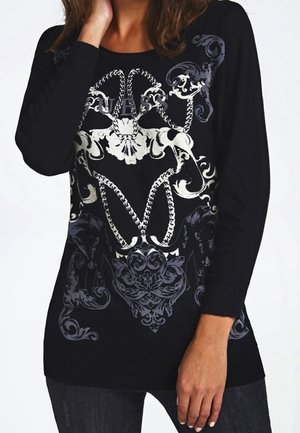 Femme portant un haut noir à manches longues avec un motif ornemental floral et en forme de corde blanc et gris complexe sur le devant.