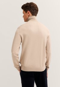 Beiger, gerippter Strickpullover mit hohem Kragen und tailliertem Saum. Weiche Textur mit nahtlosen Schultern und einem dezenten Logo-Tag an der Seite.