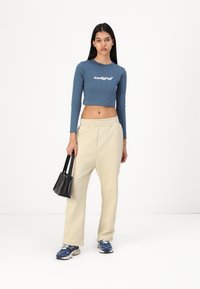 Zila garām piedurknēm cropped top ar baltu logo, krēmkrāsas sporta bikses un zilas kedas. Aksesuāri ietver mazu melnu rokassomu.
