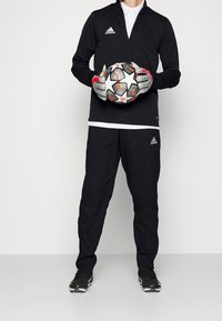 Černá atletická bunda s polozipem, černé kalhoty a rukavice. Drží míč na fotbal s hvězdným vzorem a živými akcenty.