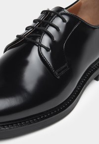 Scarpa da uomo in pelle nera con finitura lucida, caratterizzata da una punta arrotondata, design con quattro occhielli per lacci e cuciture a contrasto lungo la suola.