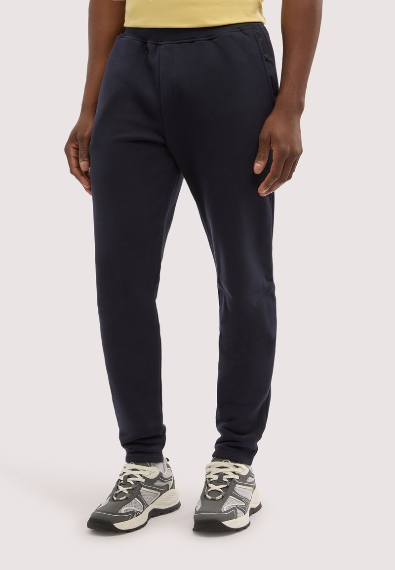 Uomo che indossa pantaloni sportivi neri a gamba stretta e scarpe da ginnastica grigie e bianche, in piedi contro uno sfondo chiaro e uniforme.