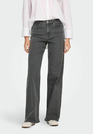Femme portant un jean noir à jambes larges, une chemise blanche boutonnée et des baskets blanches, debout devant un fond clair uni.