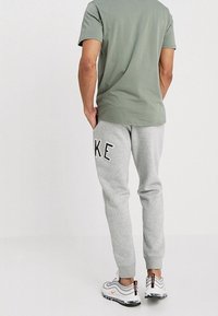 Sportovní oblečení zahrnující přiléhavé olivově zelené tričko, šedé jogger kalhoty s logem "NIKE" a šedo-bílé tenisky s oranžovými akcenty.