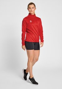 Haut de sport rouge à manches longues avec col à demi-fermeture éclair, associé à un short noir. Le haut présente un logo et des accents blancs contrastants sur les épaules.