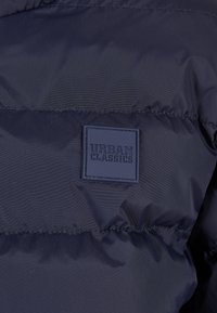 Mørkeblå pufferjakke med horisontale sømmer. Har et strukturet, firkantet stoff merke merket "URBAN CLASSICS."