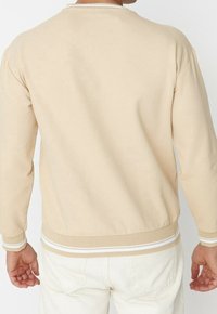 Hombre vestido con una camisa beige de manga larga con puños y dobladillo acanalados, combinada con pantalones color blanco roto, de espaldas a la cámara contra un fondo liso.