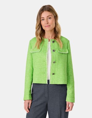 Grüne kurze Jacke aus strukturiertem Stoff, mit großen Klapptaschen, zwei Knöpfen und ausgefransten Kanten, kombiniert mit grauen Hosen.