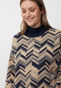 Sweter o fakturze, z wysokim kołnierzem, zdobiony geometrycznym wzorem w rybę w granatowym i beżowym kolorze. Materiał wydaje się pikowany i miękki.