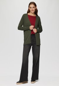 Grüne Strickjacke mit langen Ärmeln, offener Front und gerippten Bündchen, kombiniert mit einem roten Oberteil und schwarzen weit geschnittenen Jeans. Lässige Schuhe mit dicker Sohle.