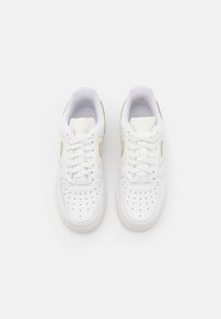 Ett par vita Nike Air-sneakers med guldaccenter, perforerat tåparti och vita snören på en helt vit bakgrund.