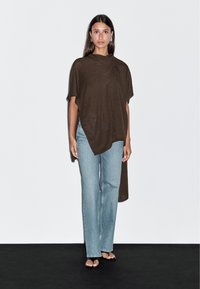 Kobieta w ciemno-brązowym asymetrycznym poncho, jasnoniebieskich dżinsach o prostym kroju, czarnych sandałach na odkrytej palcach i srebrnych kolczykach w kształcie kółek, na jednolitym tle.