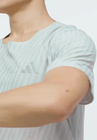 Svetlo šedé športové tričko s krátkymi rukávmi, s textúrovaným pruhovaným vzorom a decentným logom na hrudi. Materiál vyzerá priedušne.