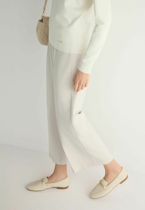 Osoba ubrana w kremowe spodnie o szerokich nogawkach, pasujące kremowe mokasyny ze złotym łańcuszkiem, biały sweter oraz beżową pikowaną torebkę na ramię.