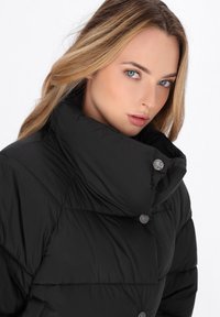 Donna con capelli biondi che indossa un giubbotto a vento nero con colletto alto e bottoni a pressione metallici su uno sfondo bianco.