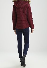 Bourgognefarvet dunjakke med pelsforet hætte og vandret quiltning. Kombineret med blå skinny jeans og sorte ankelstøvler med hæl.