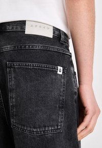 Schwarze Jeans aus Denim mit strukturiertem Finish, ausgestattet mit einem cremefarbenen Lederschild am Bund und einem kleinen weißen Etikett an der Gesäßtasche.