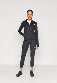 Μαύρο αθλητικό μπουφάν με φερμουάρ και κολάν, και τα δύο από ελαστικό ύφασμα, με λογότυπα στο στήθος και στη γάμπα. Φορεμένα με μαύρα αθλητικά παπούτσια.