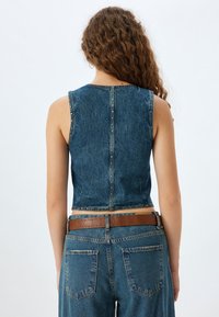 Gilet di denim tagliato in blu scuro con una finitura testurizzata, caratterizzato da borchie metalliche lungo la cucitura posteriore. Abbinato a jeans a vita alta.