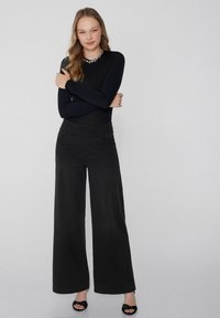 Blusa negra de manga larga con un cuello decorativo de cadena plateada, combinada con pantalones negros de pierna ancha que cuentan con bolsillos frontales.