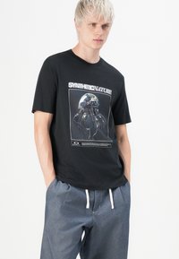 Black cotton t-shirt featuring a graphic print of a jellyfish and earth, with "SYNTHETIC NATURE" text above. Worn with gray casual pants.