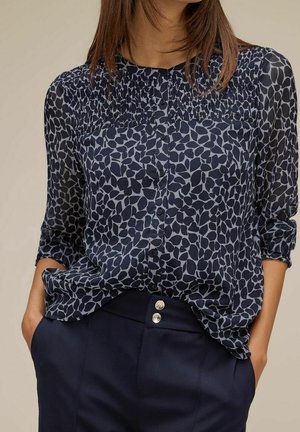 Blouse bleu marine à motif de feuilles, avec un empiècement froncé, une patte de boutonnage à l'avant et des manches trois-quarts, associée à un pantalon foncé.