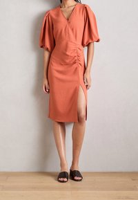 Robe couleur rouille, longueur genoux avec des manches bouffantes, des détails froncés sur les côtés et une fente haute sur la cuisse. Associée à des sandales glissantes noires.