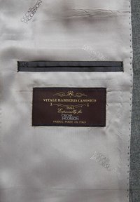 Etichetta di Vitale Barberis Canonico attaccata a un tessuto per abito grigio. Presenta cuciture, un'etichetta marrone scuro e dettagli in filo metallico.