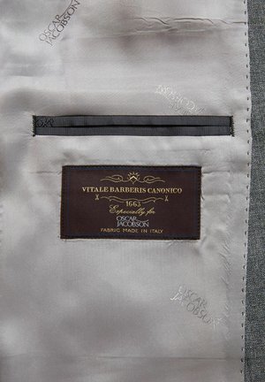 Label van Vitale Barberis Canonico bevestigd aan grijze suitstof. Bevat stiksel, een donkerbruine label en details in metalen draad.