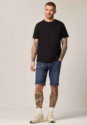 Homme aux bras et jambes tatoués portant un t-shirt noir, un short en jean, des chaussettes blanches et des baskets beiges, debout devant un fond uni.