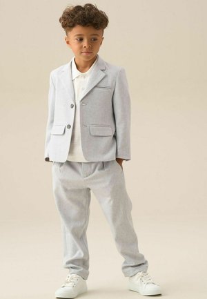 Jeune garçon aux cheveux bouclés portant un costume gris clair, un polo blanc et des baskets blanches, debout les mains dans les poches.