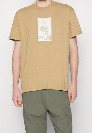 T-shirt beige à manches courtes avec un graphisme d’une grue de chantier et un texte, porté avec un pantalon cargo vert olive par une personne debout.