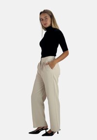 Pantaloni beige a vita alta con pinces e tasche, abbinati a un dolcevita nero aderente. La modella è in profilo e indossa tacchi neri.