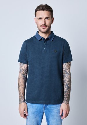 Camisa polo de rayas en azul marino con un cuello en contraste, presenta un parche con el logo, mangas cortas y una textura ligera. Combinada con jeans de un azul claro.