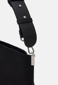 Cinghia di una borsa in pelle nera con gancio metallico argentato attaccato al bordo di una borsa in camoscio o pelle nera su uno sfondo bianco semplice.