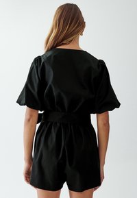 Mono negro de manga corta con cintura ceñida y cinturón, que presenta una silueta relajada y textura suave, mostrado desde la parte trasera.