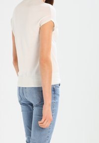 Svetle béžový svetr s krátkými rukávy a texturovaným povrchem, přiléhavý střih a žebrovaný lem, kombinovaný se světle modrými džíny.