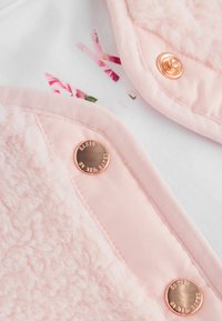 Gaiši rozā jaka ar pūkainu tekstūru vienā pusē, gludu audumu otrā pusē, rotāta ar rozā zelta spiedpogām un ziedu iekšējo druku.