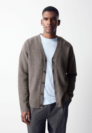 Man wearing a brown buttoned cardigan over a light blue shirt and gray pants, standing with one hand in his pocket against a plain background.