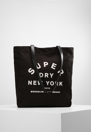 Czarna torba na ramię z dwoma czarnymi uchwytami, biały tekst "Super Dry New York Tokyo Brooklyn Bronx", wyświetlony na białej powierzchni.