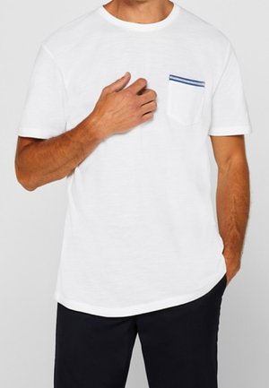 Homme portant un t-shirt blanc uni à manches courtes avec une petite poche poitrine comportant deux bandes bleues, associé à un pantalon noir.