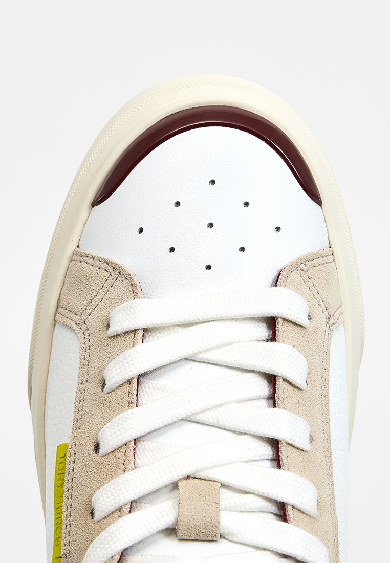 Weiße Leder-Sneaker mit beigefarbenen Wildlederakzenten, rotem Gummitrim, perforierter Vorderseite und weißen Schnürsenkeln. Verfügt über ein gelbes Logodetail an der Seite.