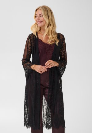 Femme souriante aux cheveux blonds portant un cardigan en dentelle noire par-dessus un haut violet foncé bordé de dentelle et un pantalon assorti, debout devant un fond uni.