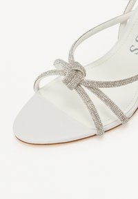 White strappy sandal featuring a smooth leather upper, embellished with a glittering crystal knot detail and a flat sole.
