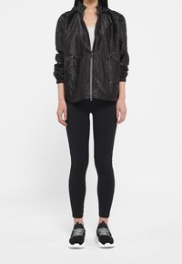 Kobieta ubrana w błyszczącą czarną kurtkę na zamek, dopasowane czarne legginsy i czarne sneakersy z białymi podeszwami i wzorzystymi sznurowadłami, stojąca na białym tle.