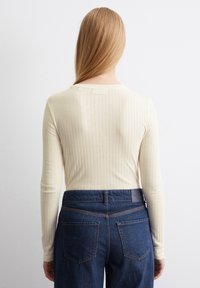 Krämfärgad ribbad långärmad tröja tillsammans med mörkblå högmidjade jeans, med bakfickor och en synlig midjebandsetikett.