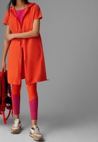 Cappotto con zip arancione e cappuccio sopra una maglietta rosa, abbinato a leggings arancioni e rosa, e sneakers beige. Zaino rosso nelle vicinanze.