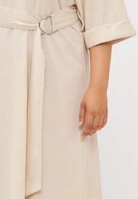 A close-up of a hand gripping a belt on a light beige garment, showcasing a relaxed style with rolled-up sleeves.
